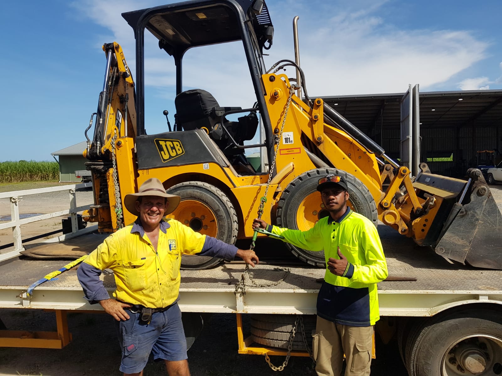 Two students in front of a backhoe having just completed their backhoe ticket.  One does a thumbs up.